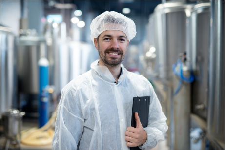 A man wearing a white safety suit and hairnet standing in a facility with a clipboard while smiling at the camera